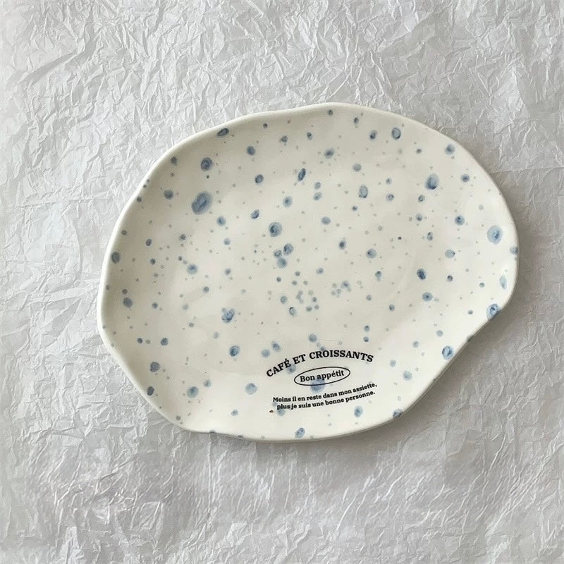 An irregularly shaped ceramic plate with a white glaze and blue speckled pattern, featuring the text 'CAFÉ ET CRÔISSANTS Bon appétit' printed on the bottom, set against a crumpled white paper background.