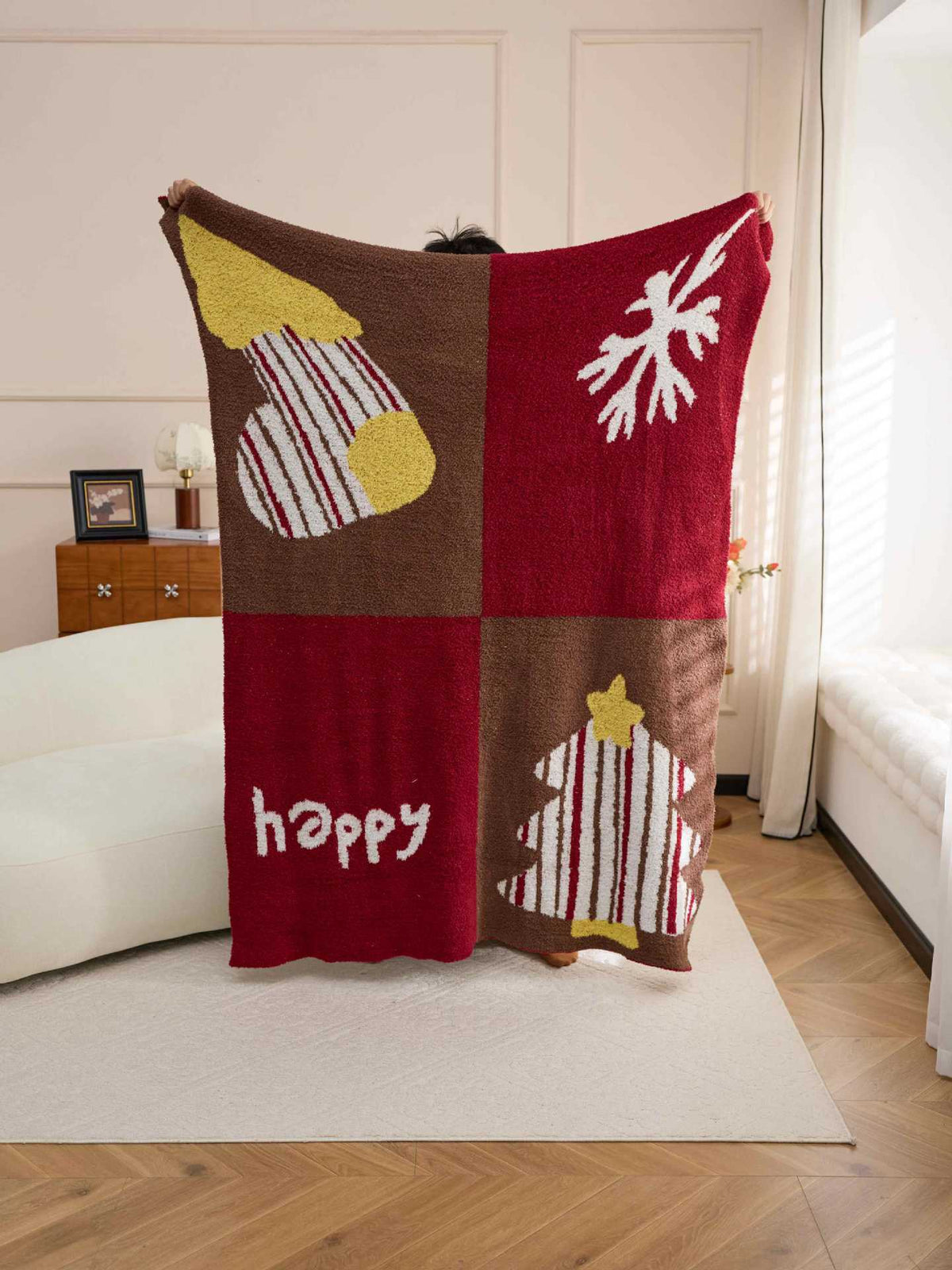 A person holds up a large red and brown holiday-themed blanket in a bright living room. The blanket has a four-quadrant design with Christmas tree, snowflake, striped ornament, and 'happy' motifs in red, brown, yellow, and white.