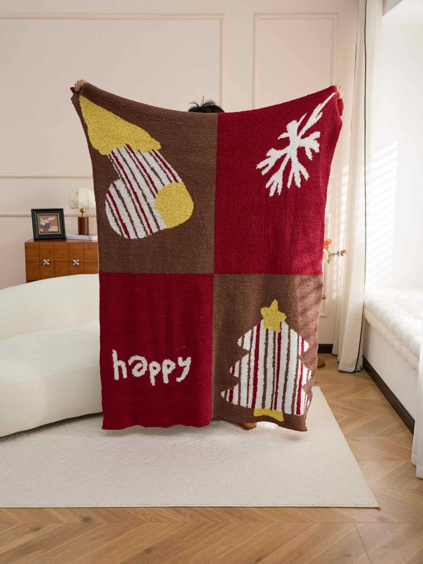 A person holds up a large red and brown holiday-themed blanket in a bright living room. The blanket has a four-quadrant design with Christmas tree, snowflake, striped ornament, and 'happy' motifs in red, brown, yellow, and white.