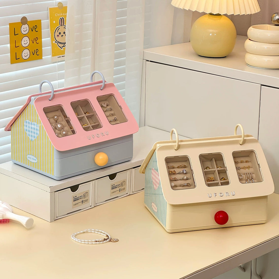 Two small jewelry boxes with compartments on a surface, one pink and green, the other pink and blue, with a book titled 'Two Thousand & Twenty Three' in the background.