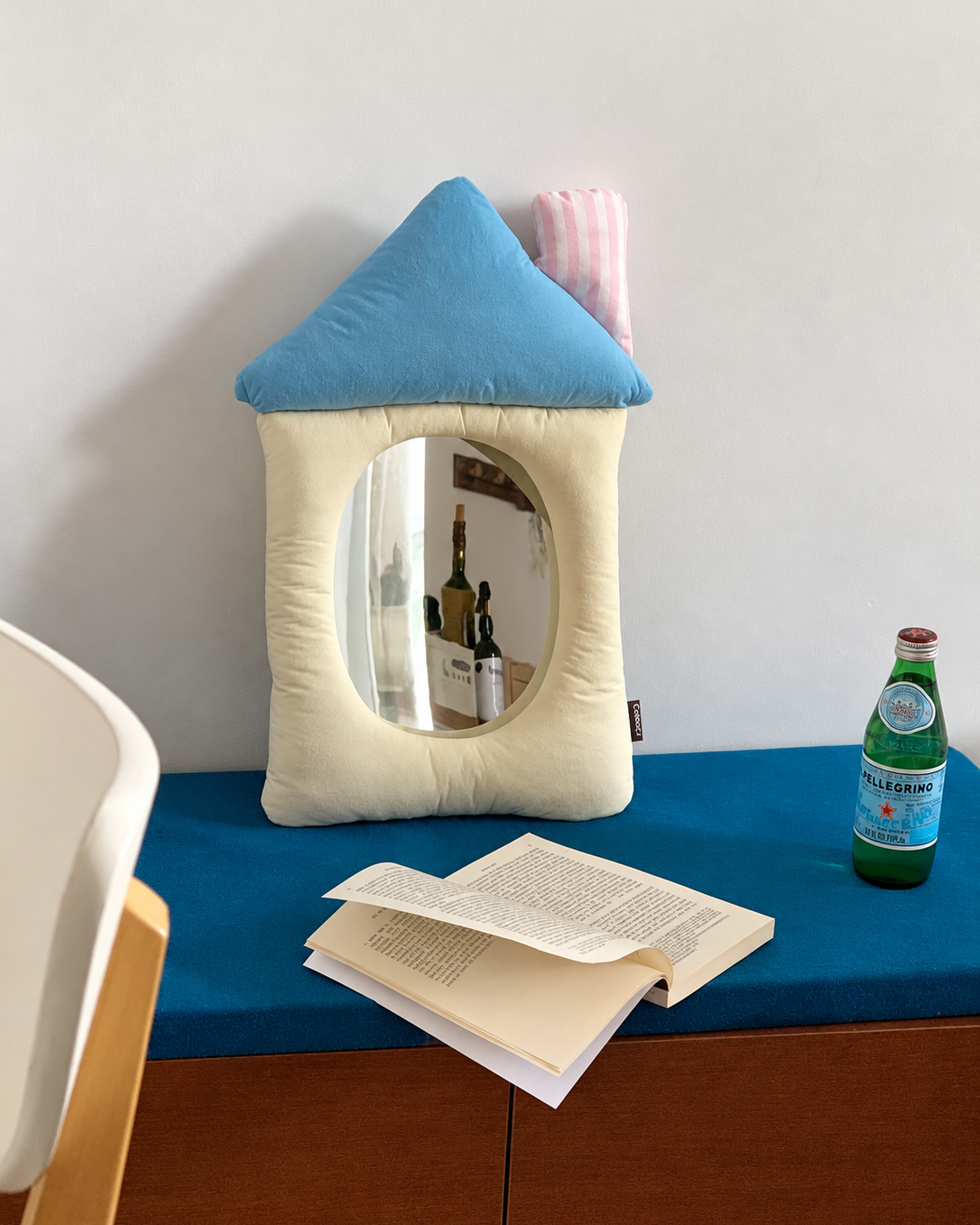 A house-shaped cushion mirror with a blue roof and cream body, set on a blue surface with an open book and a green bottle nearby.