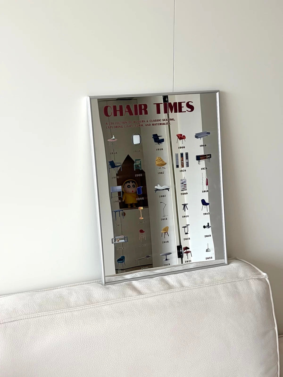 A rectangular wall mirror with a thin silver frame, leaning against a white wall on a white upholstered chair. The mirror reflects a room interior and features a printed vintage-style chart titled 'CHAIR TIMES' with small illustrations of different chairs and text labels.