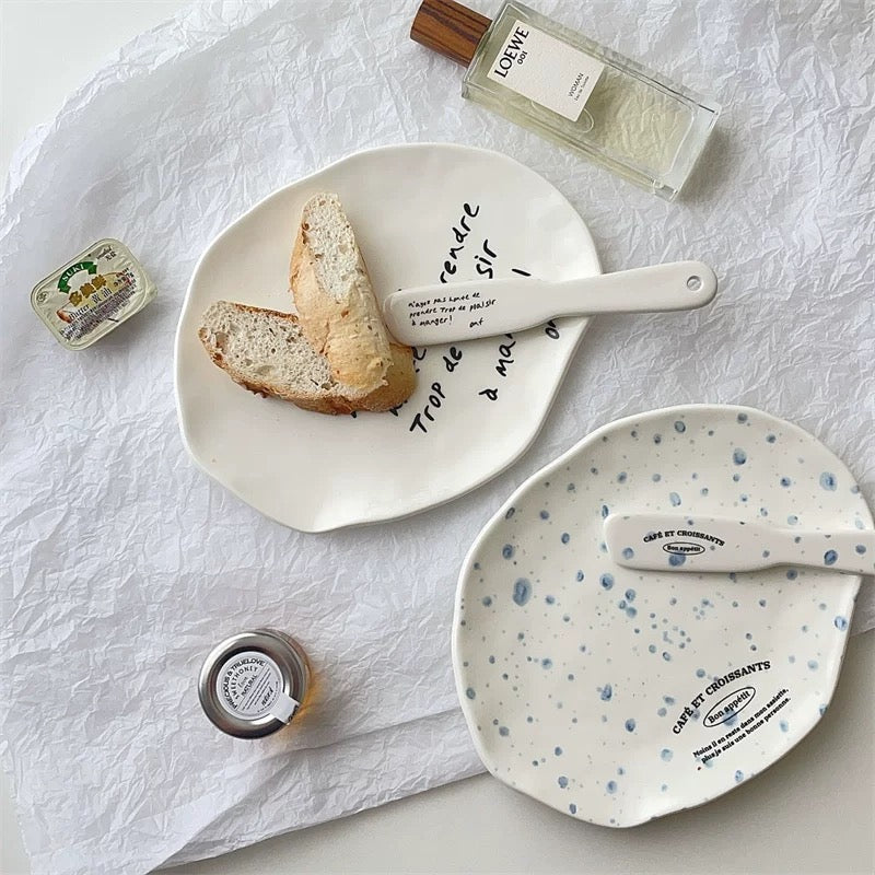 Two ceramic plates with spoons, styled on a crumpled white paper background with bread, a bottle of perfume, and small jars as props.