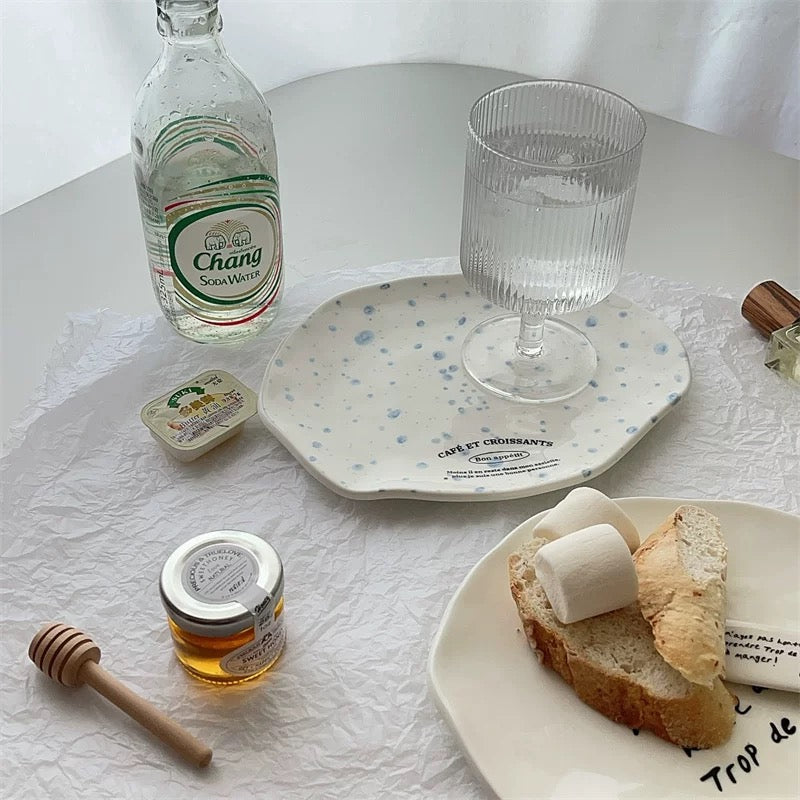 A styled tabletop scene with a white ceramic plate featuring blue speckles and the text 'CRAFTED ENGRAAVEMENTS' holding a clear ribbed glass. A bottle of Chang Suan Mei, a small jar of honey, a wooden honey dipper, marshmallows on bread, and other breakfast items are arranged on a white textured cloth.