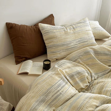 A cozy bed styled with a cream and beige striped duvet cover and matching pillowcases, accented by a solid brown pillow and an open book on the bedside table.