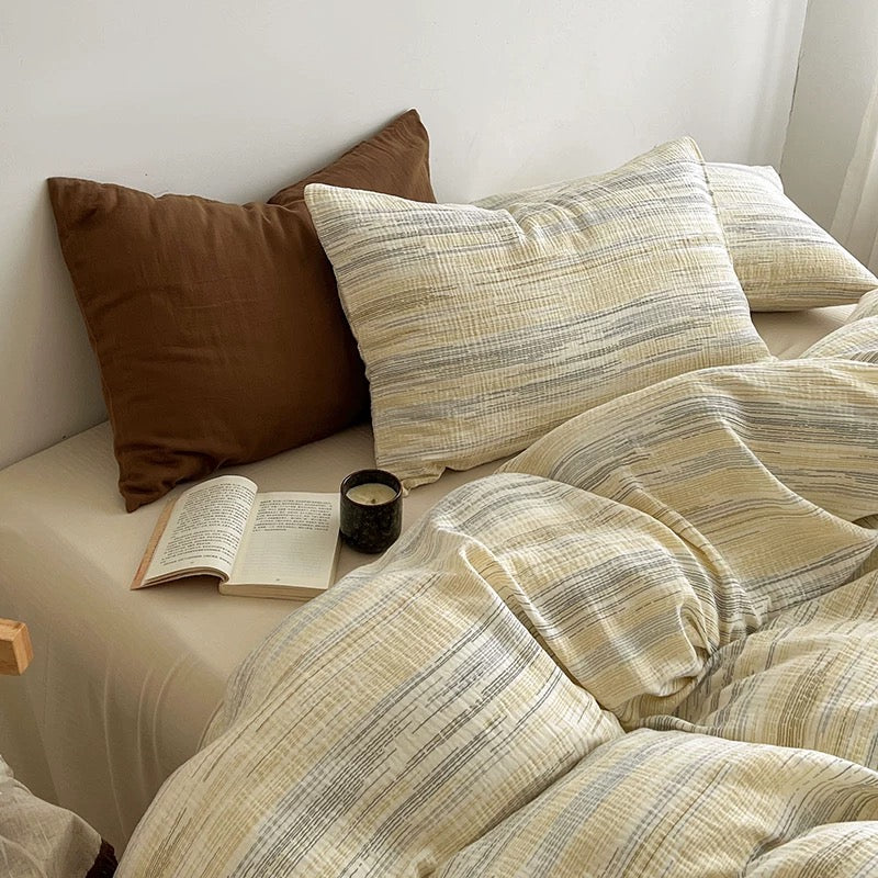 A cozy bed styled with a cream and beige striped duvet cover and matching pillowcases, accented by a solid brown pillow and an open book on the bedside table.