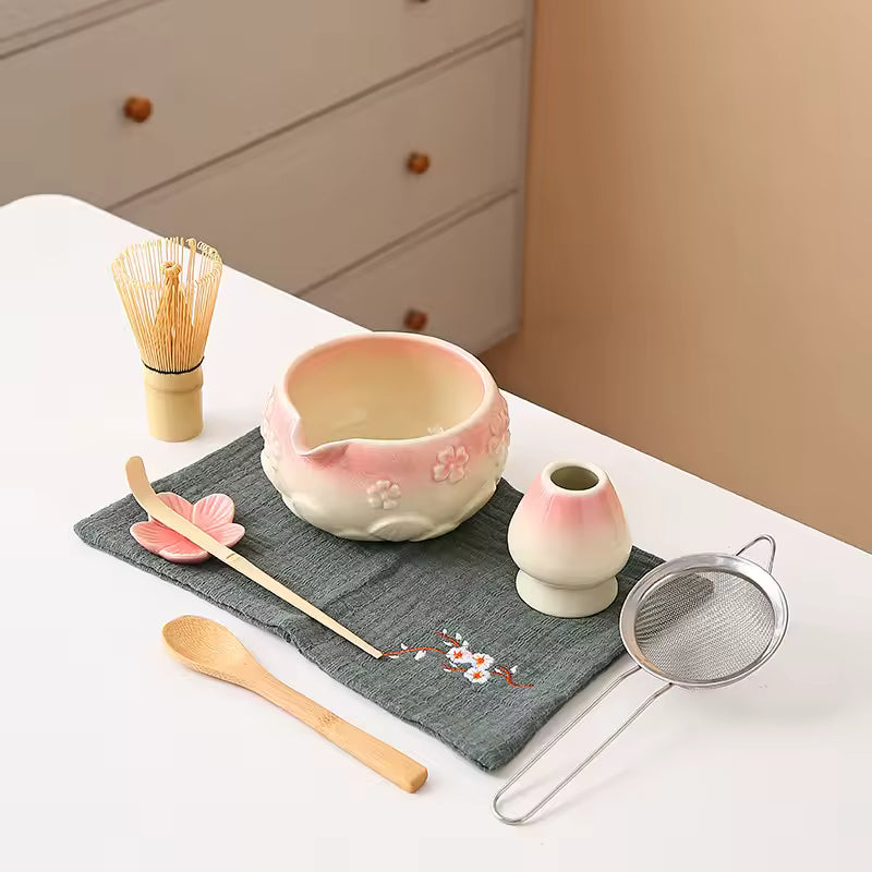 Ceramic tea set with a pink bowl, whisk, spoon, and strainer on a gray cloth.