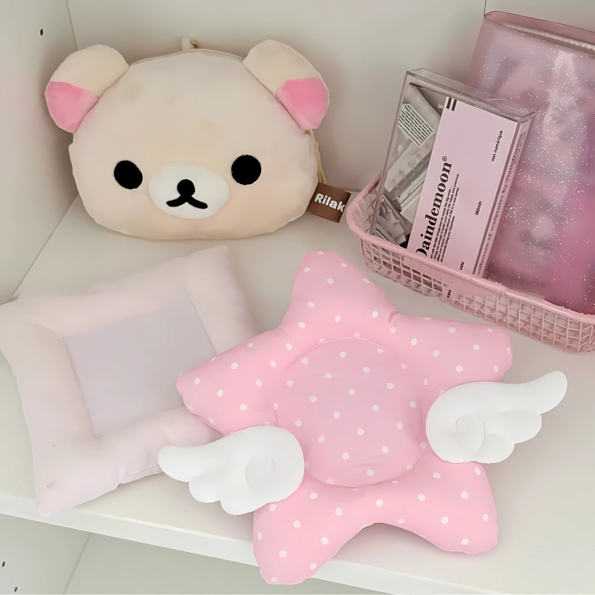 Pink star-shaped pillow with angel wings on a shelf with a plush bear toy and a pink box.
