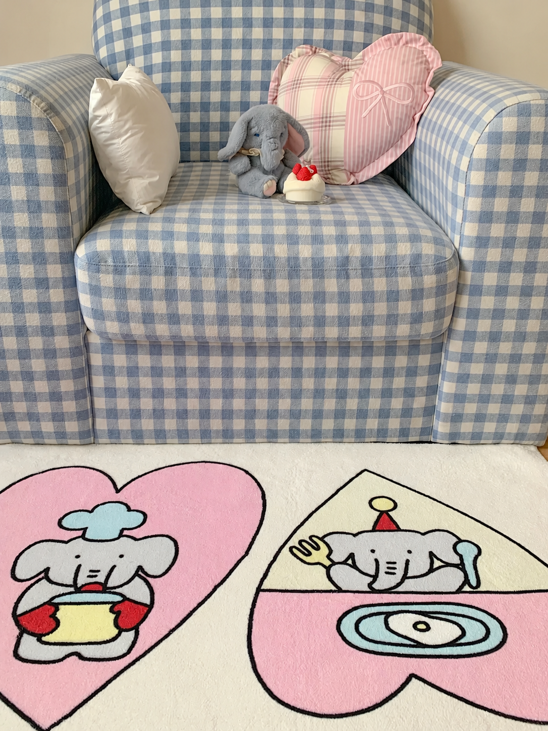 A cutecore rug with two pink hearts and cooking elephants lies beside a white shelf on a cream rectangular base.