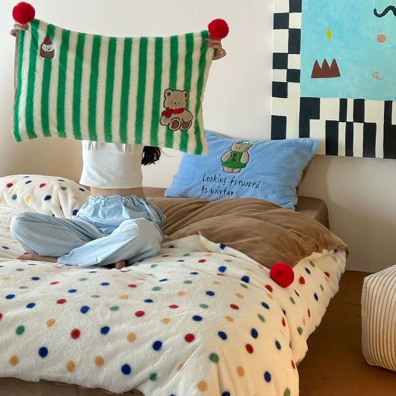 Person holding a green and white striped pillow with red pom-poms in a bedroom setting.