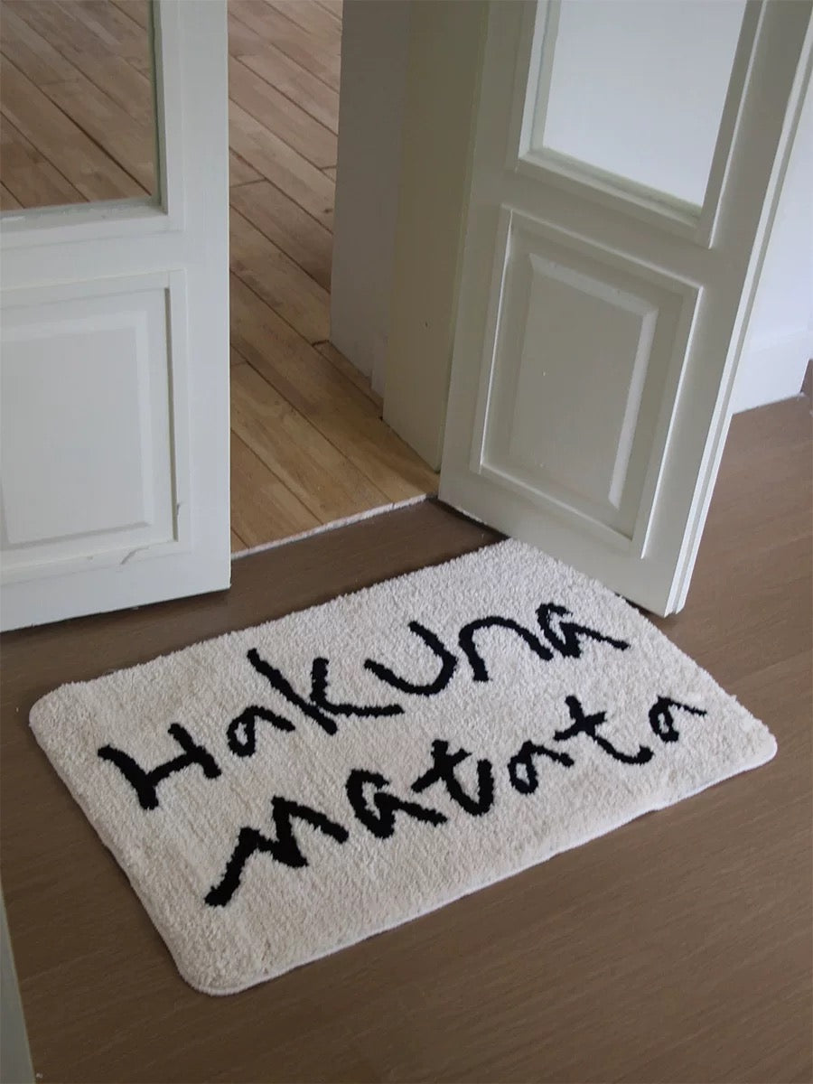 A Korean bath mat in white displays black Hakuna Matata lettering beside an open indoor door.