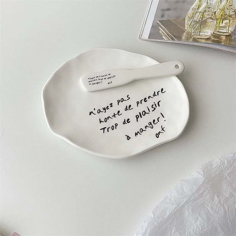 A white ceramic plate with an organic, hand-drawn shape, featuring black handwritten French text inside and a matching white ceramic spoon with printed text resting on top. The plate is photographed on a clean, light gray surface with a magazine corner visible in the background.