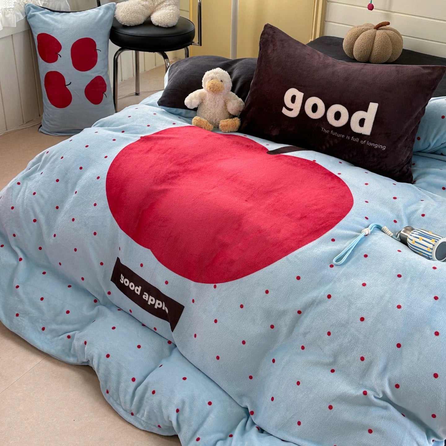 Blue bedspread with red apple design and 'good app' text, surrounded by pillows and a plush toy.