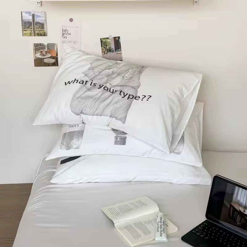 A stack of white pillows on a bed, with the top pillow printed in black text reading 'what is your type??' over a gray-toned abstract design. The bed is styled with a book and a laptop, against a cream wall with small decorative prints pinned to it.
