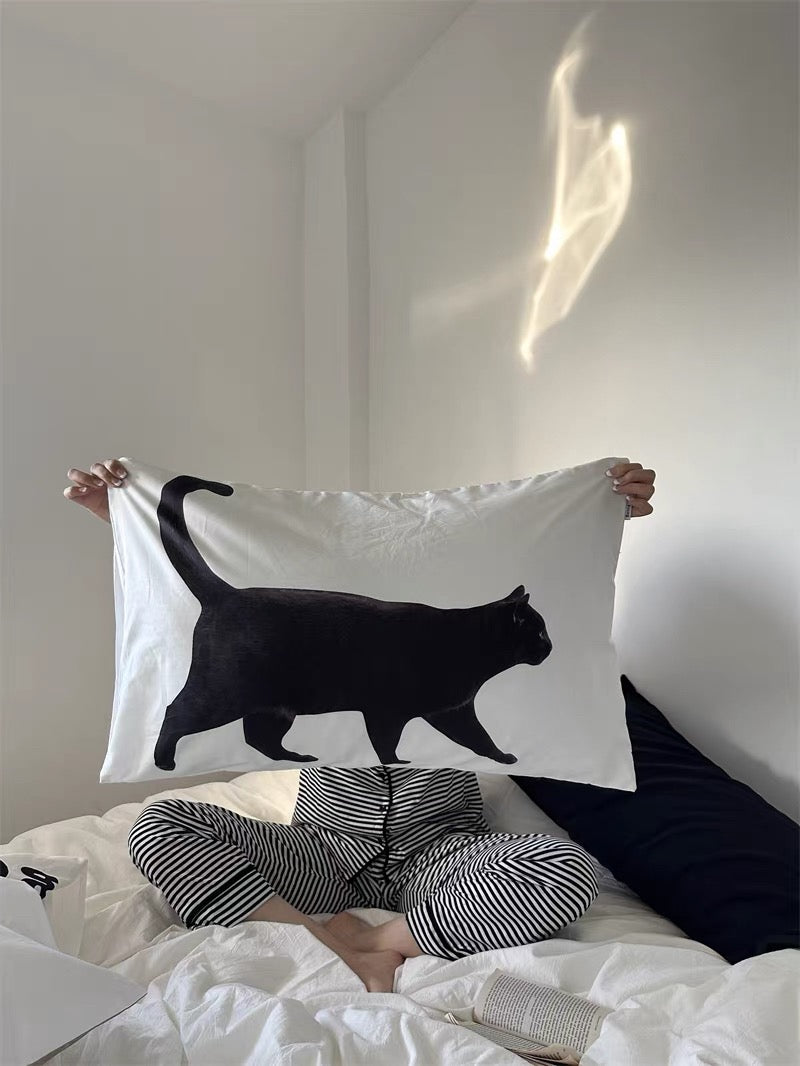 Person holding a white pillowcase with a black cat design on a bed.