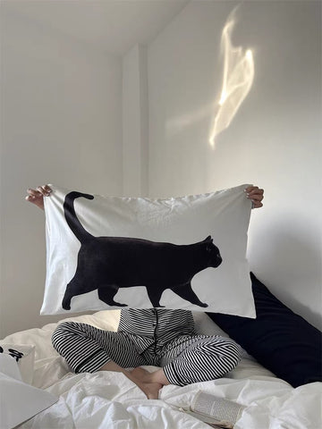Person holding a white pillowcase with a black cat design on a bed.