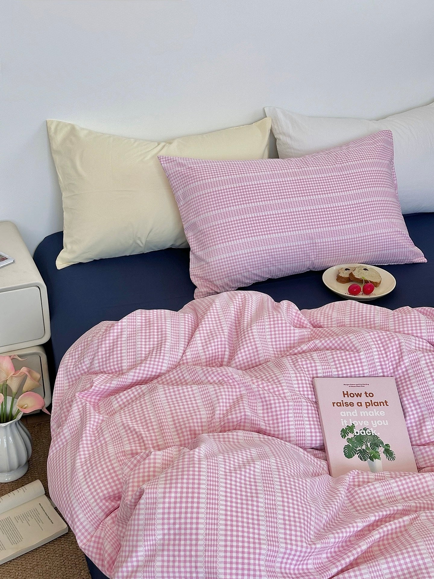 Bed with pink checkered duvet and pillows, nightstand with books and flowers.