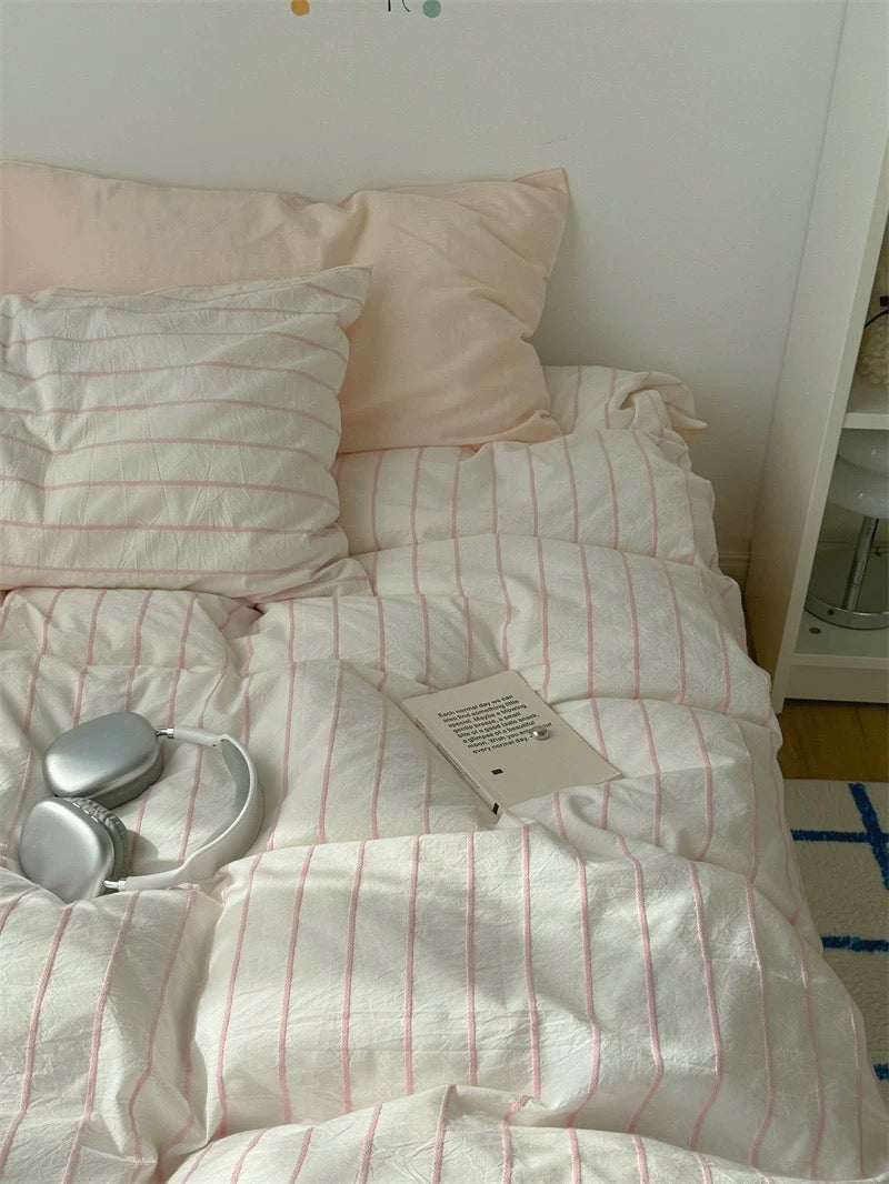 Bed with white and pink striped bedding, pink pillows, and a small book on a nightstand.