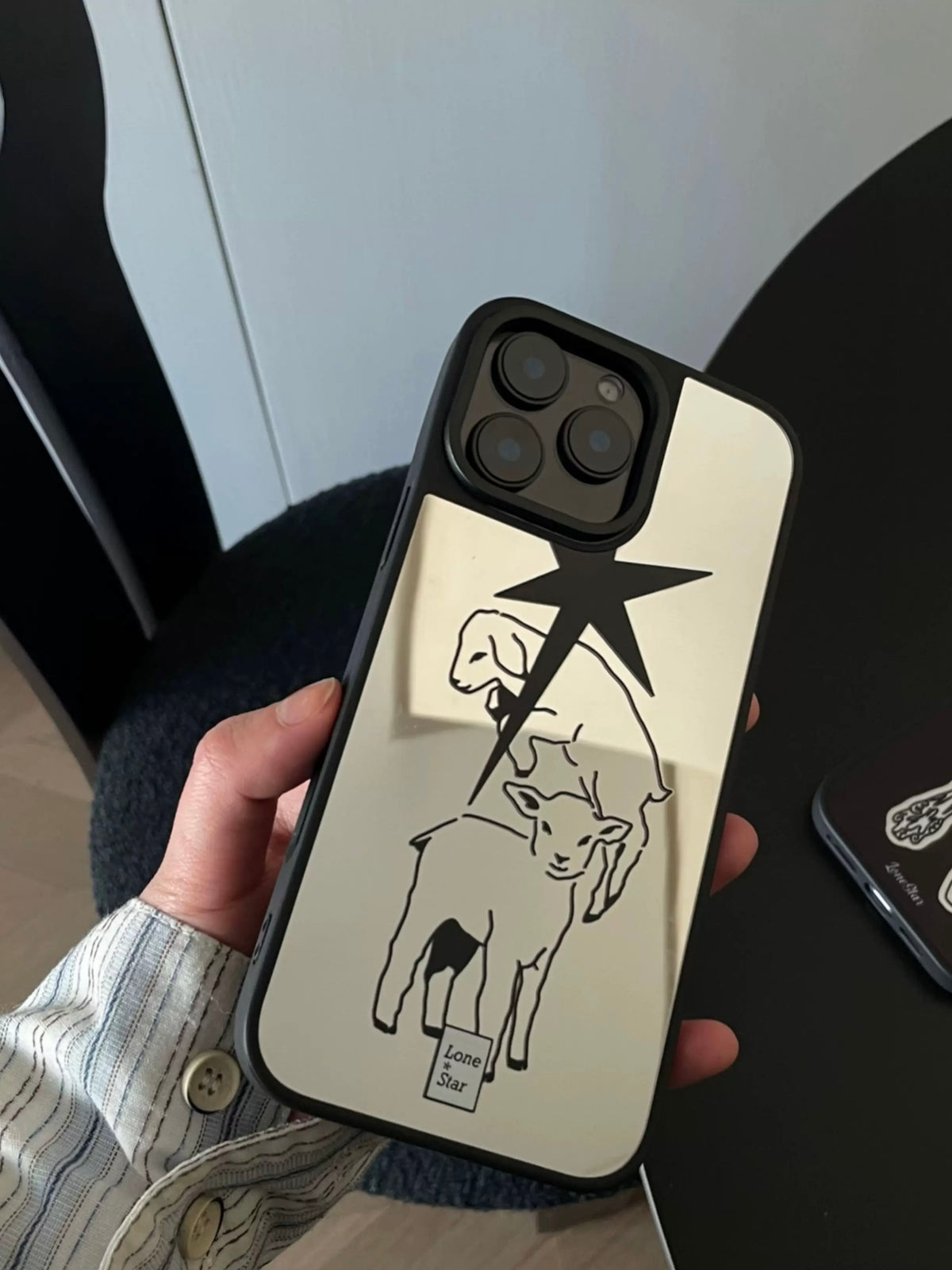 A lamb phone case is held upright, showing a reflective silver back, black pointed star and black edging.