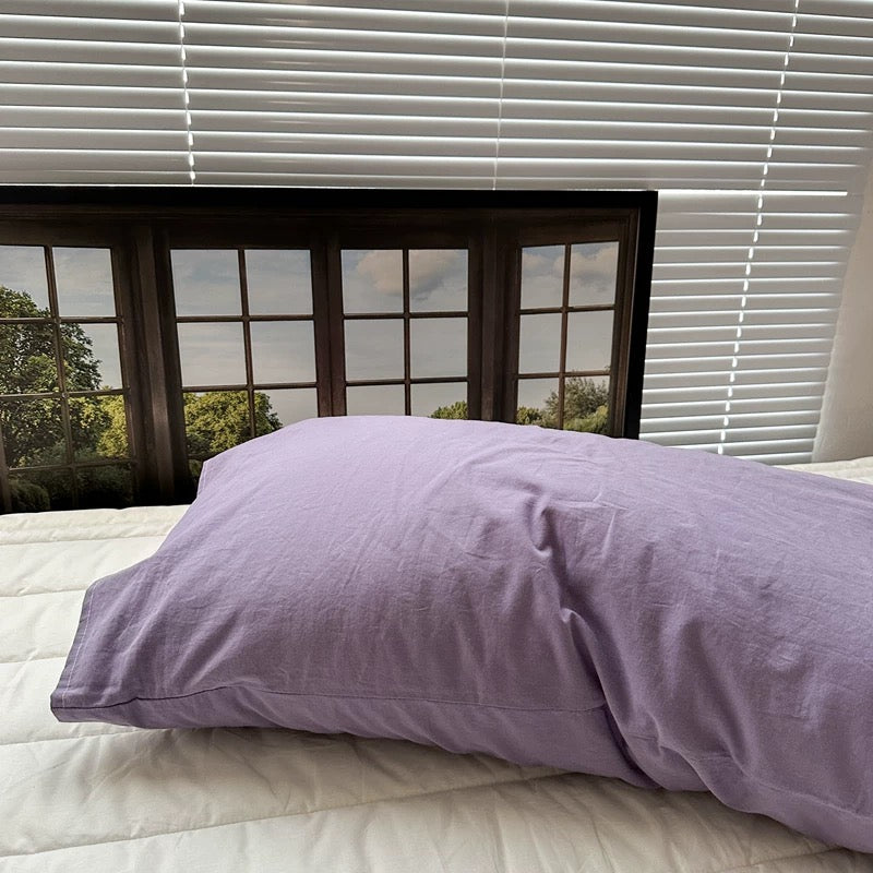 A lavender pillow cover rests on a white bed beneath a framed window print.