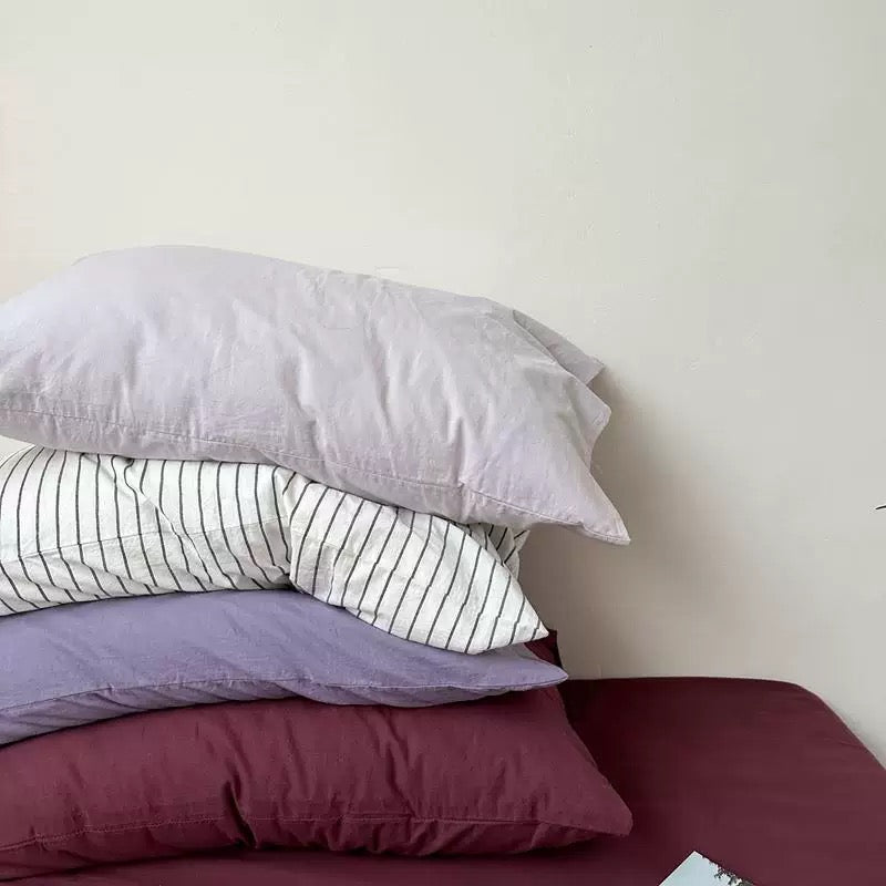 A lavender pillowcase is stacked with plum, pale gray, and white striped pillowcases on a white bed.