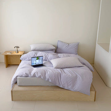A minimalist platform bed with light wood flooring, dressed in a lavender and white striped bedding set with matching pillows, a small wooden bedside table, and a laptop on the bed.