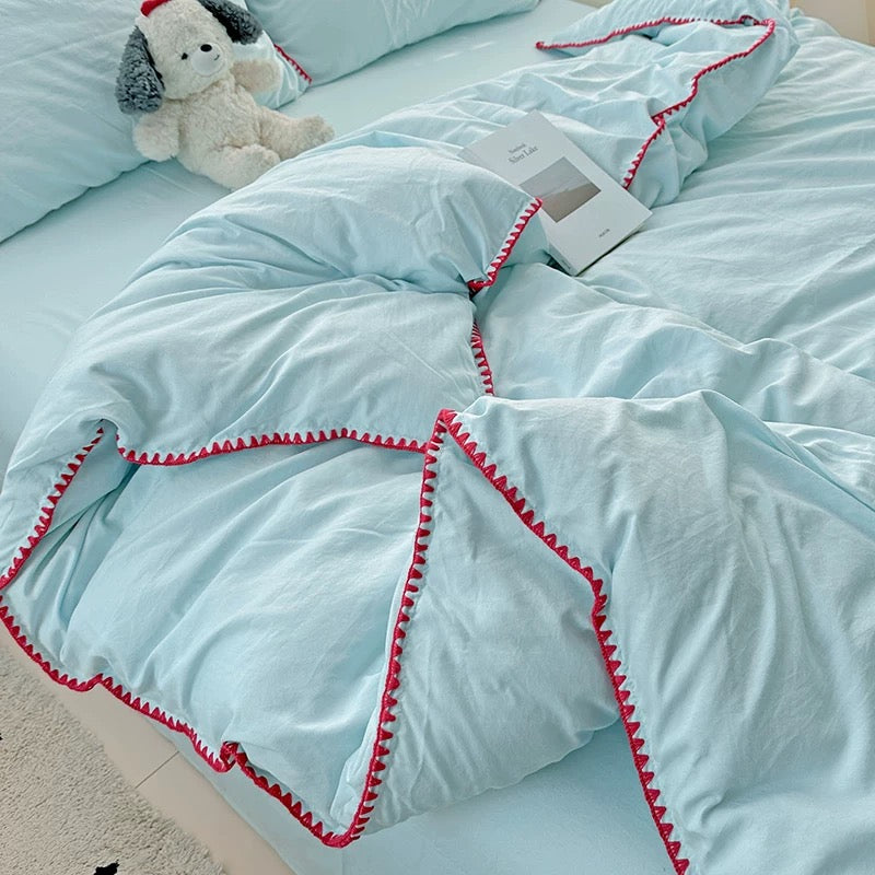 Light blue comforter with red trim on a bed, featuring a plush toy.