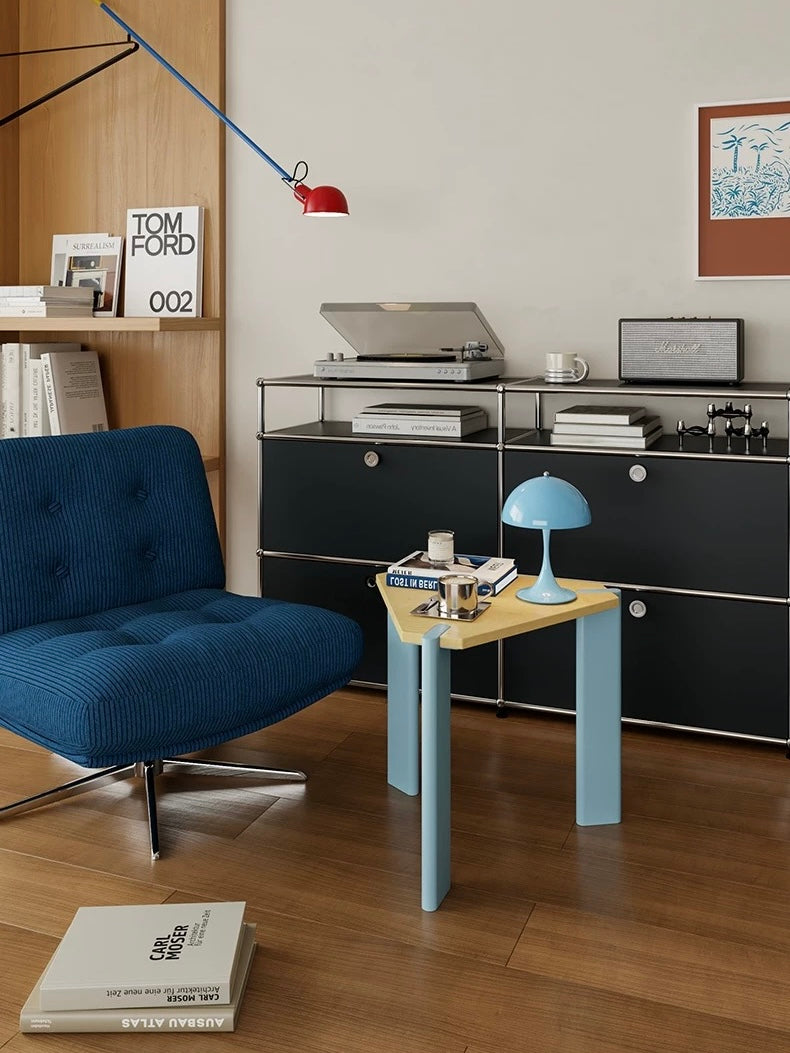 Light blue side table with a triangular wood top holding a blue lamp and small objects beside a blue chair.