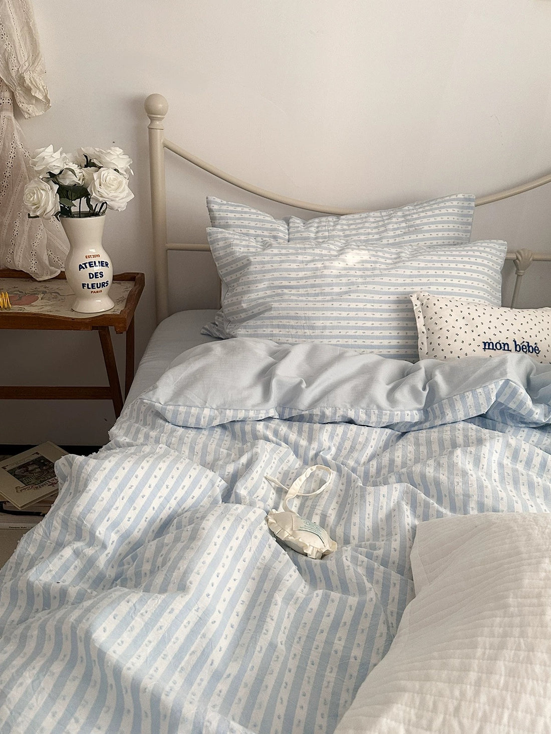 A neatly made bed with light blue and white striped bedding, including a duvet cover and matching pillowcases, styled in a cozy bedroom setting with a wooden side table, white flowers, and a small decorative pillow.