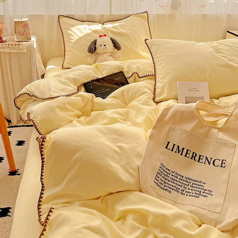 A soft cream-yellow bedding set with matching pillows and a duvet cover, styled on a bed in a cozy bedroom. The bedding has decorative brown cord trim, and a small plush dog toy rests on one pillow.