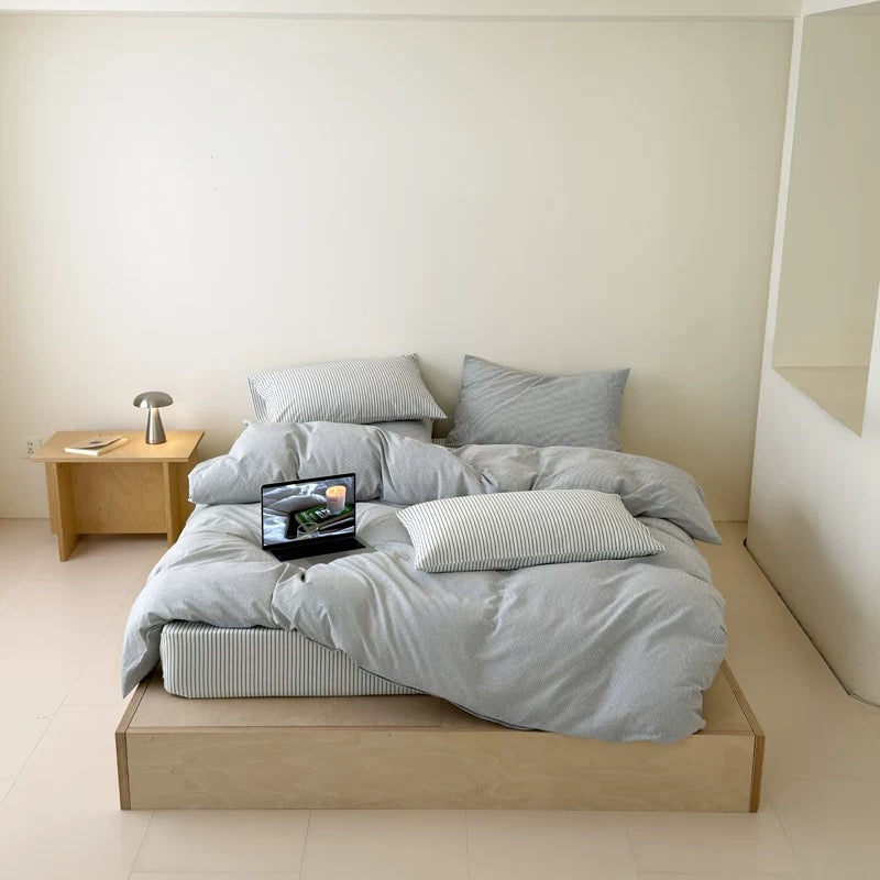 A minimalist bedroom scene featuring a light gray and white striped bedding set on a low wooden platform bed. The bedding includes a duvet cover, multiple pillows, and a matching striped mattress protector.