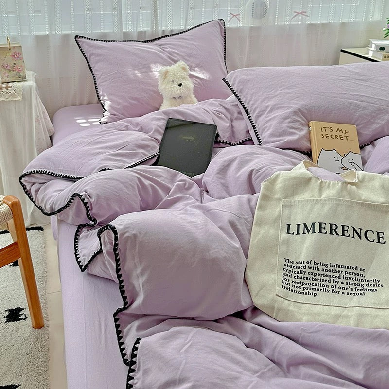 A lavender bedding set with black pom-pom trim, styled on a bed with matching pillows, a small plush toy, books, and a cream canvas tote bag in a bright bedroom setting.