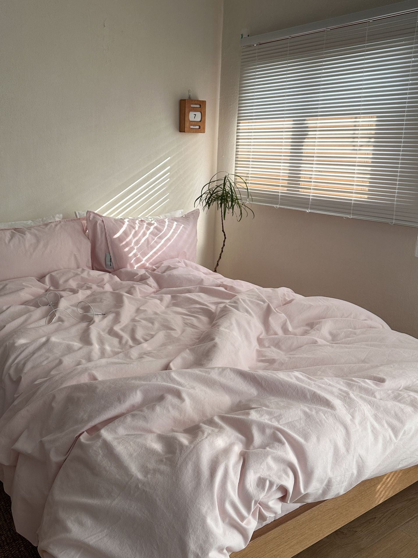 A bed dressed in light blush pink bedding, including a duvet cover and matching pillows, styled in a minimalist bedroom with a window, blinds, and a small plant.