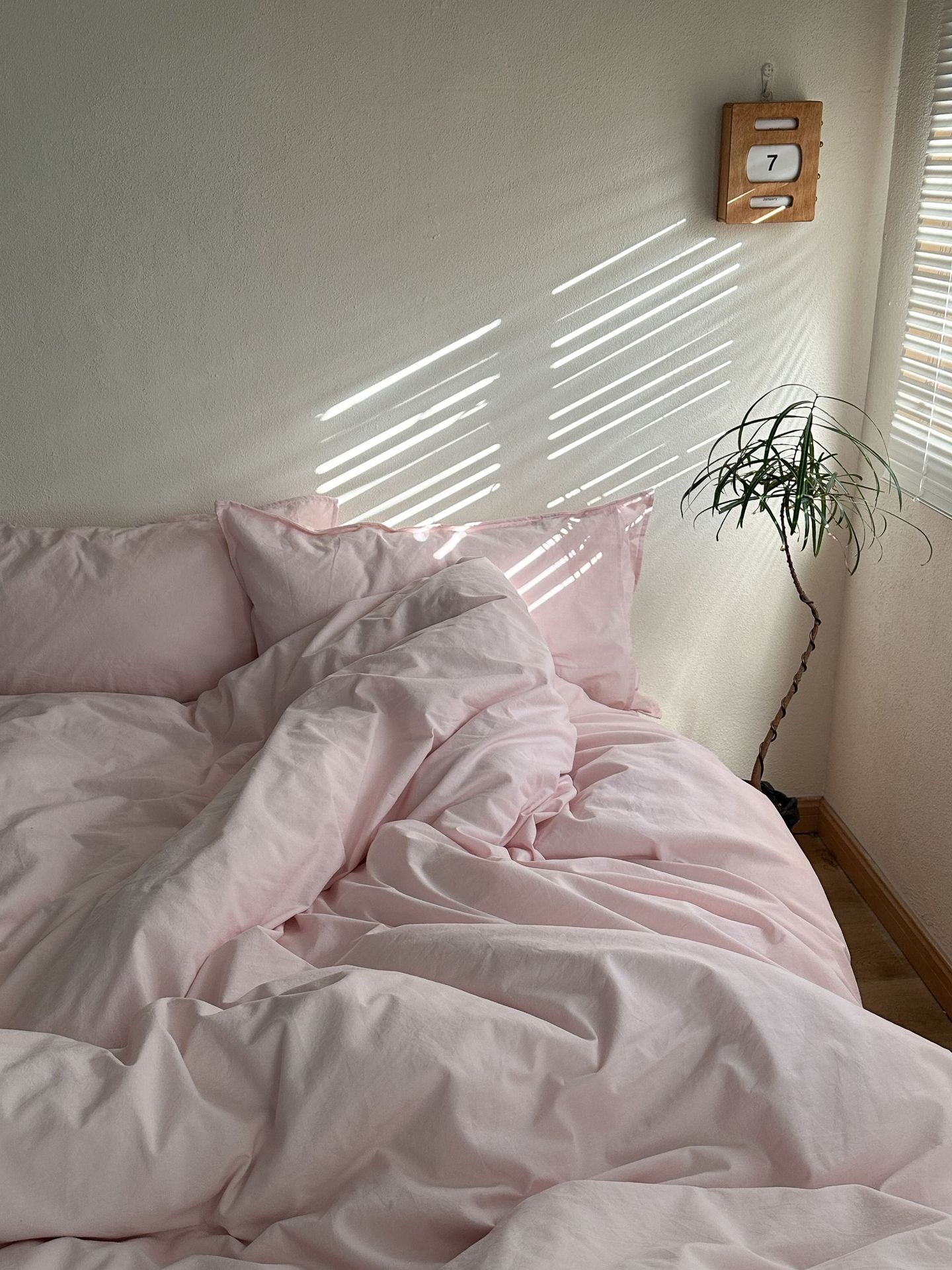 A bed dressed in soft blush pink bedding, including a duvet cover and matching pillows, styled in a sunlit bedroom with window blinds casting diagonal shadows on the wall.