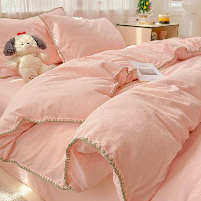 A pink bedding set with matching duvet cover and pillowcases, styled on a bed in a softly lit bedroom. The bedding has delicate light green zigzag trim along the edges, and a small plush dog toy sits on the bed.