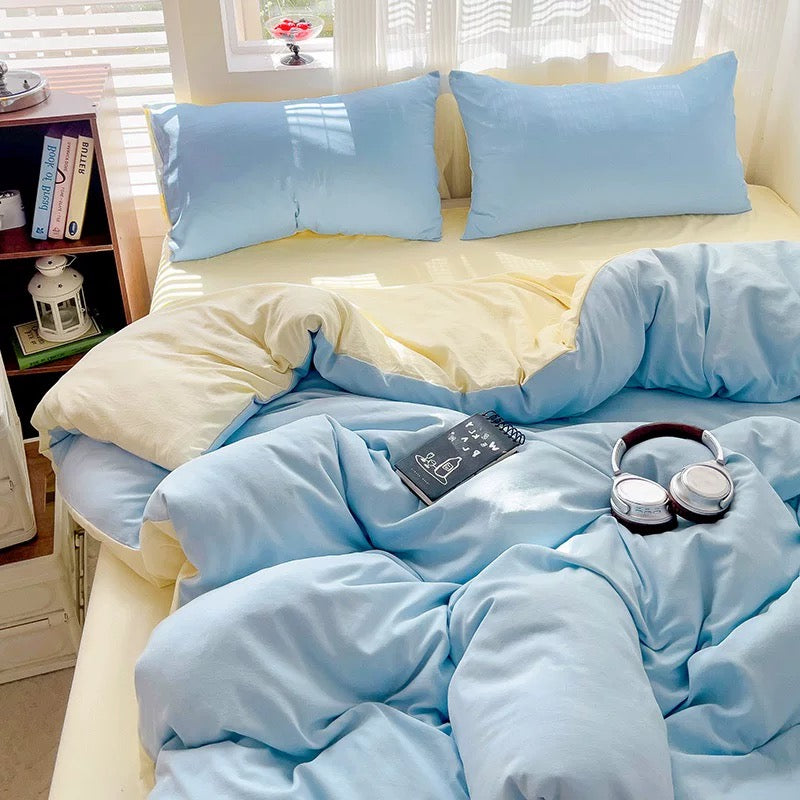 Light blue bedding set on a bed in a bright room with windows and shelves.