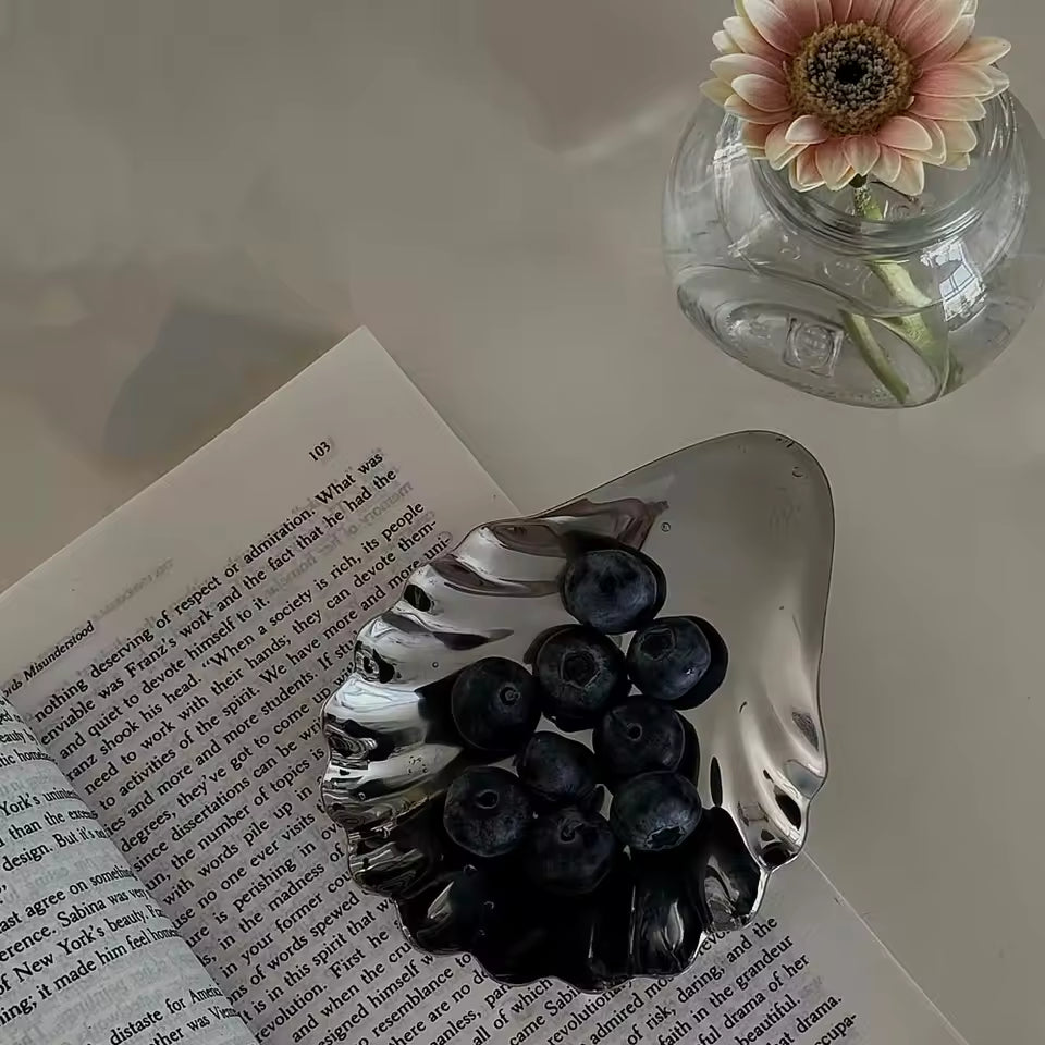 Silver dish with blueberries on an open book next to a pink flower in a glass vase.