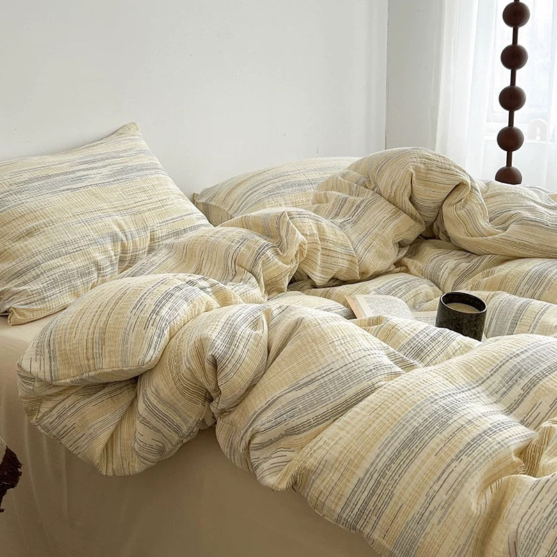A bed made with a textured, crinkled cotton bedding set in beige and muted gray stripes, styled with rumpled duvet cover and pillows in a bright, minimalist bedroom.