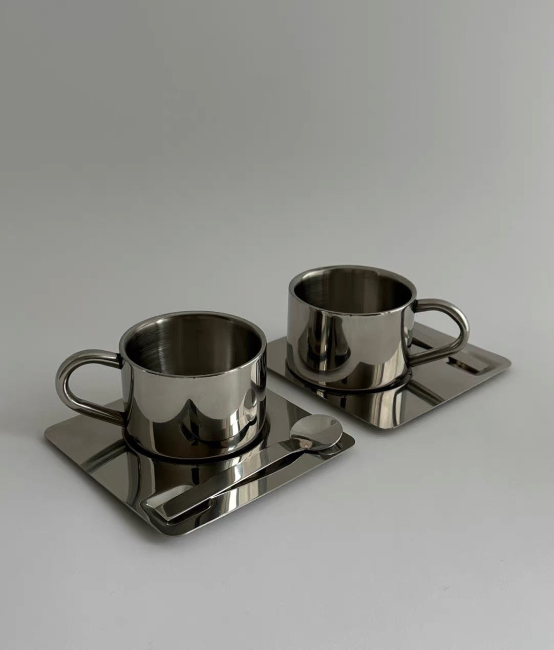 Two metallic cups with saucers on a reflective surface