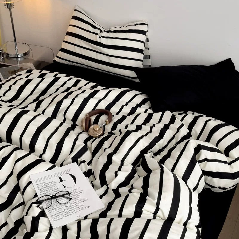 Black and white striped bedding set with matching pillowcases on a bed, styled with a black pillow, a pair of over-ear headphones, glasses, and a magazine in a bedroom setting.