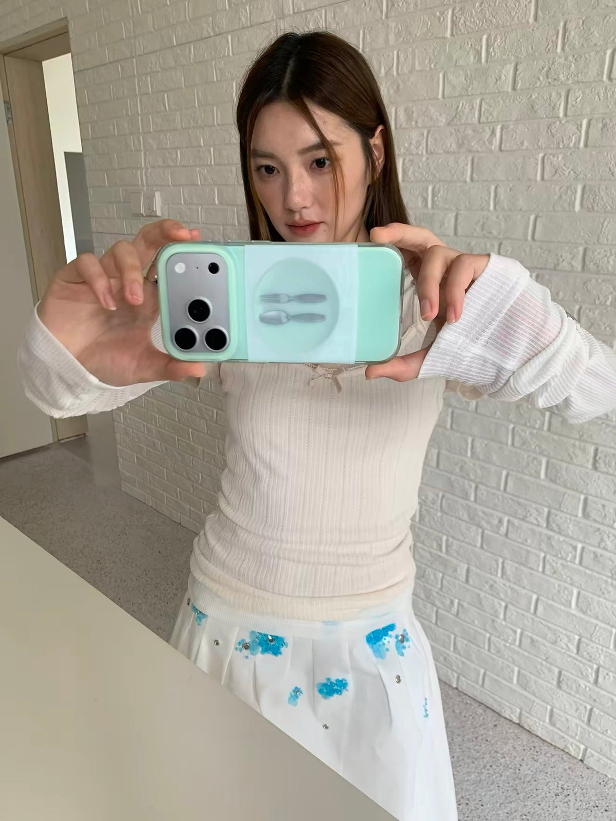 A mint green plate phone case in a wider mirror selfie, held sideways with its illustrated back visible.