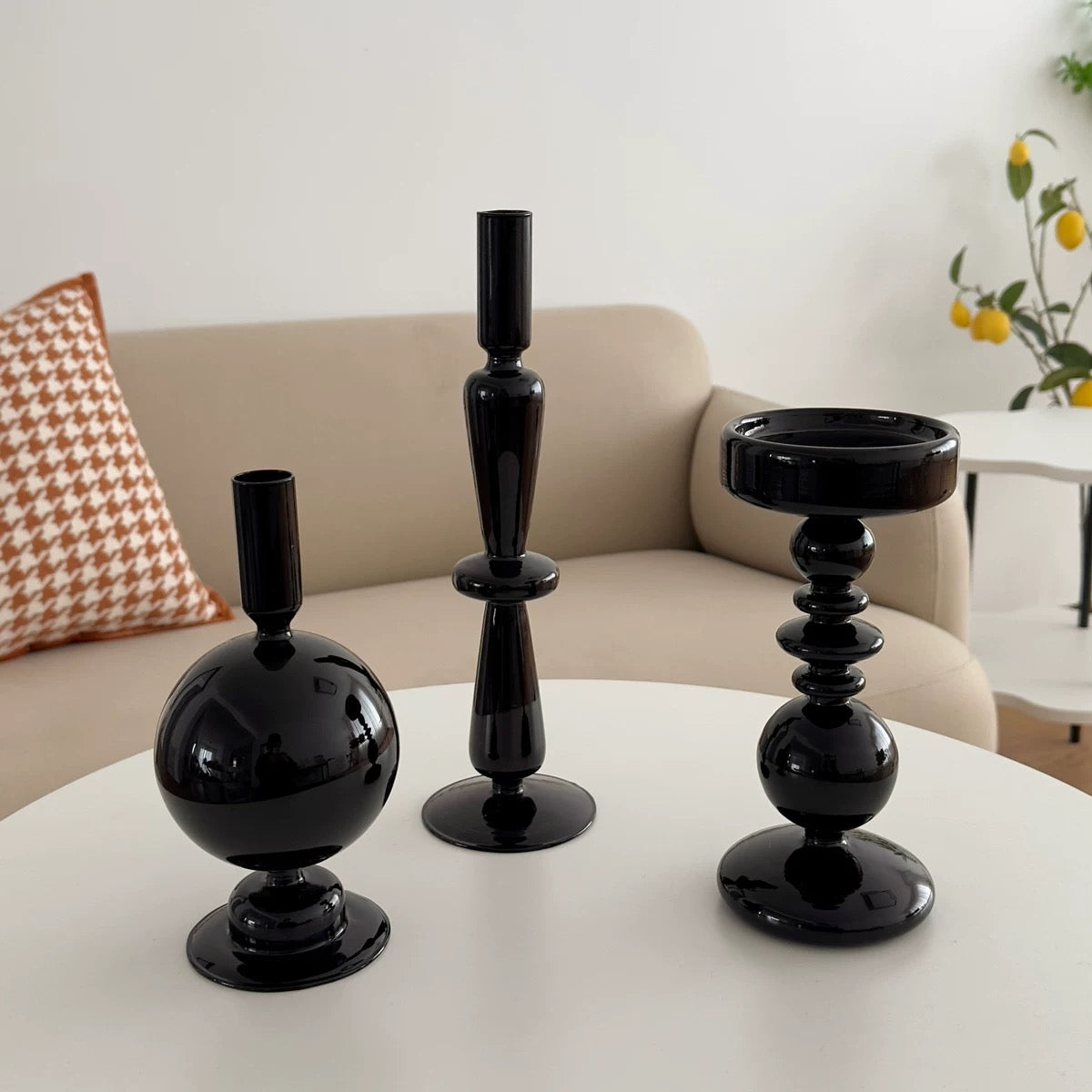 Three black candle holders on a white table with a beige sofa and decorative pillows in the background.