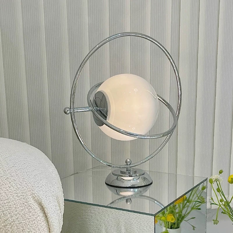 A modern table lamp with a glowing white spherical shade and a chrome-finished orbiting metal frame, displayed on a mirrored glass side table.