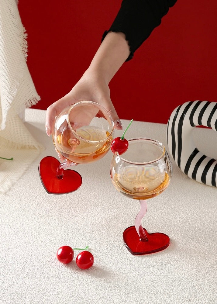 Two glasses with heart-shaped bases and red cherries on a white surface with a red background.