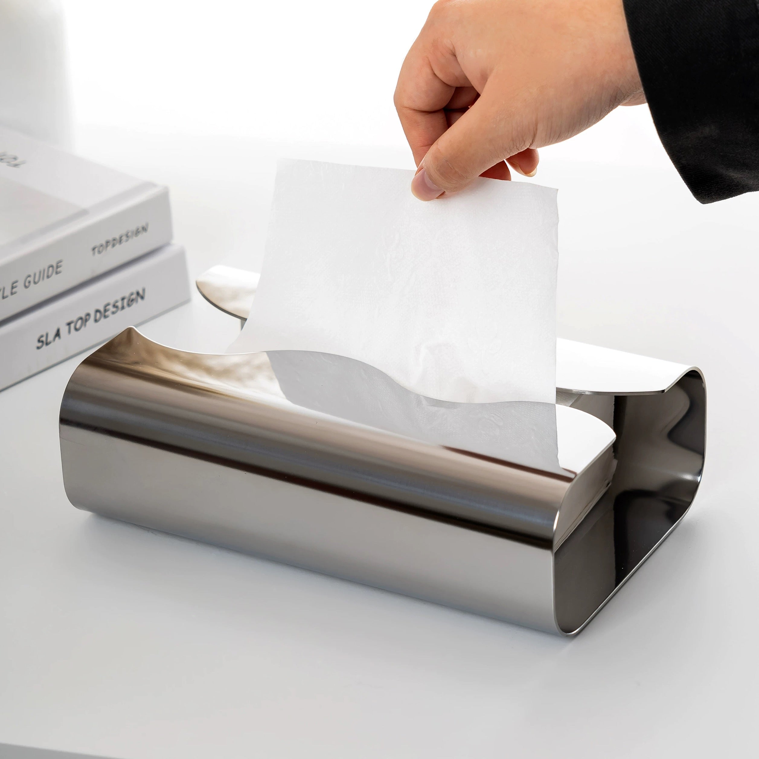 Hand pulling a sheet of paper from a metallic paper towel holder on a white surface.
