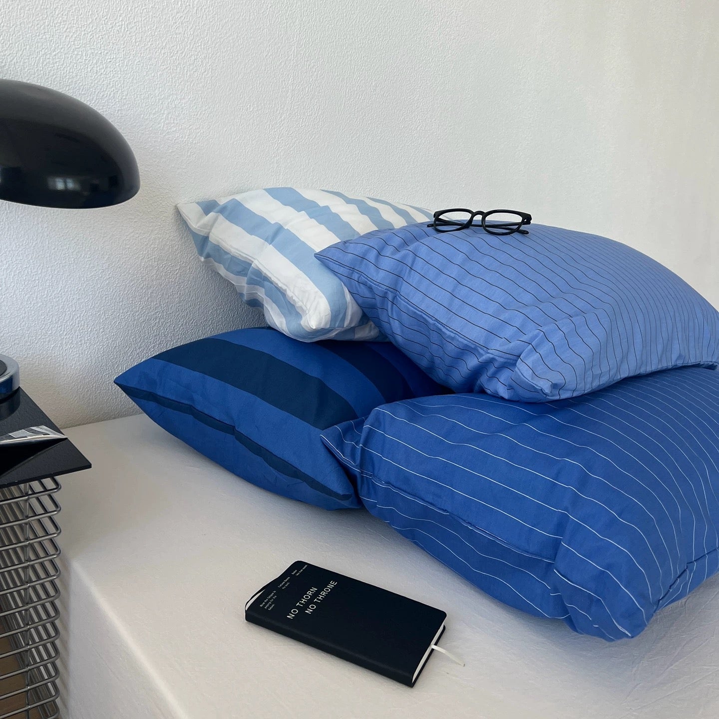 Three blue patterned pillows arranged on a white bedside table next to a black lamp, with a pair of black eyeglasses resting on the top pillow.