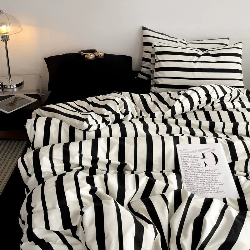 A bed dressed in a black and white striped bedding set, including a duvet cover and two matching pillowcases, styled in a bedroom setting with a bedside lamp, black pillow, and magazine.