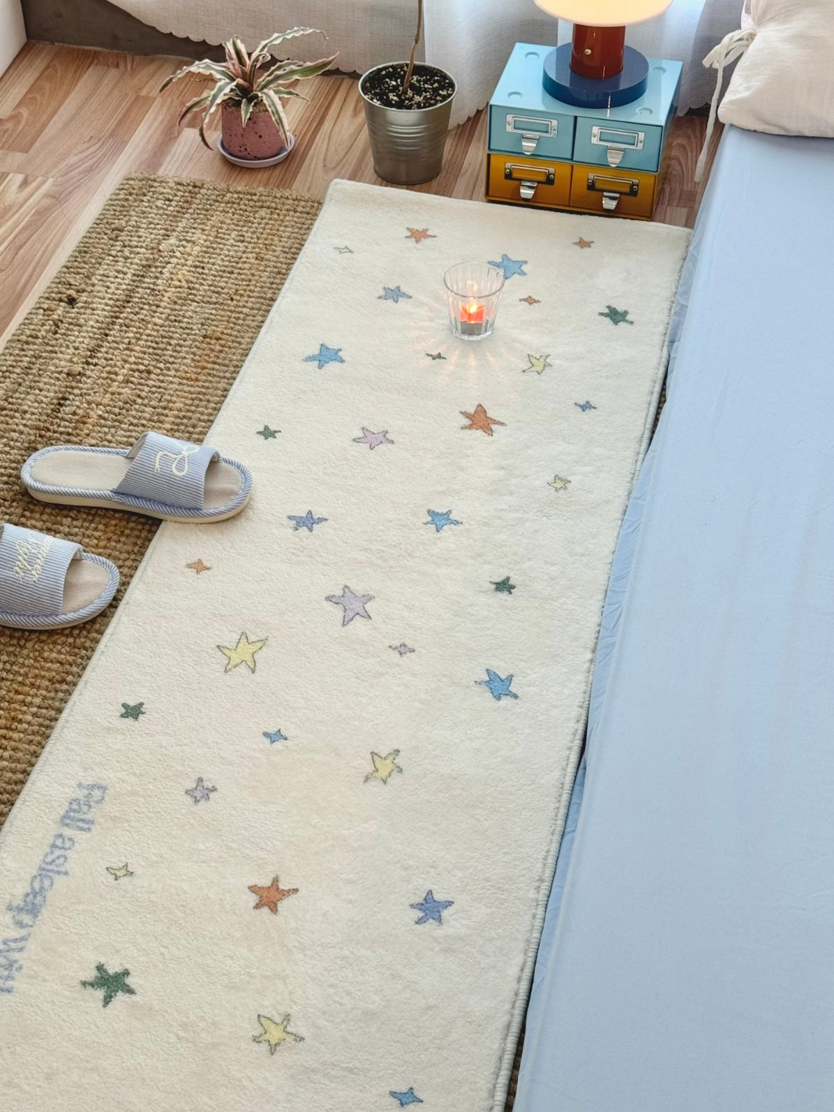 A multicolor star runner rug lies along a blue bed, with scattered stars and a glass near the far end.