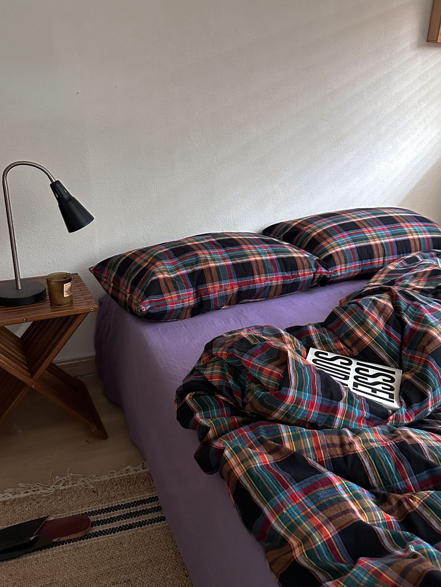 A bed dressed in a purple sheet and matching plaid bedding, with two pillows and a rumpled duvet in dark navy, red, green, and beige tartan. A wooden side table with a black lamp sits beside the bed against an off-white wall.