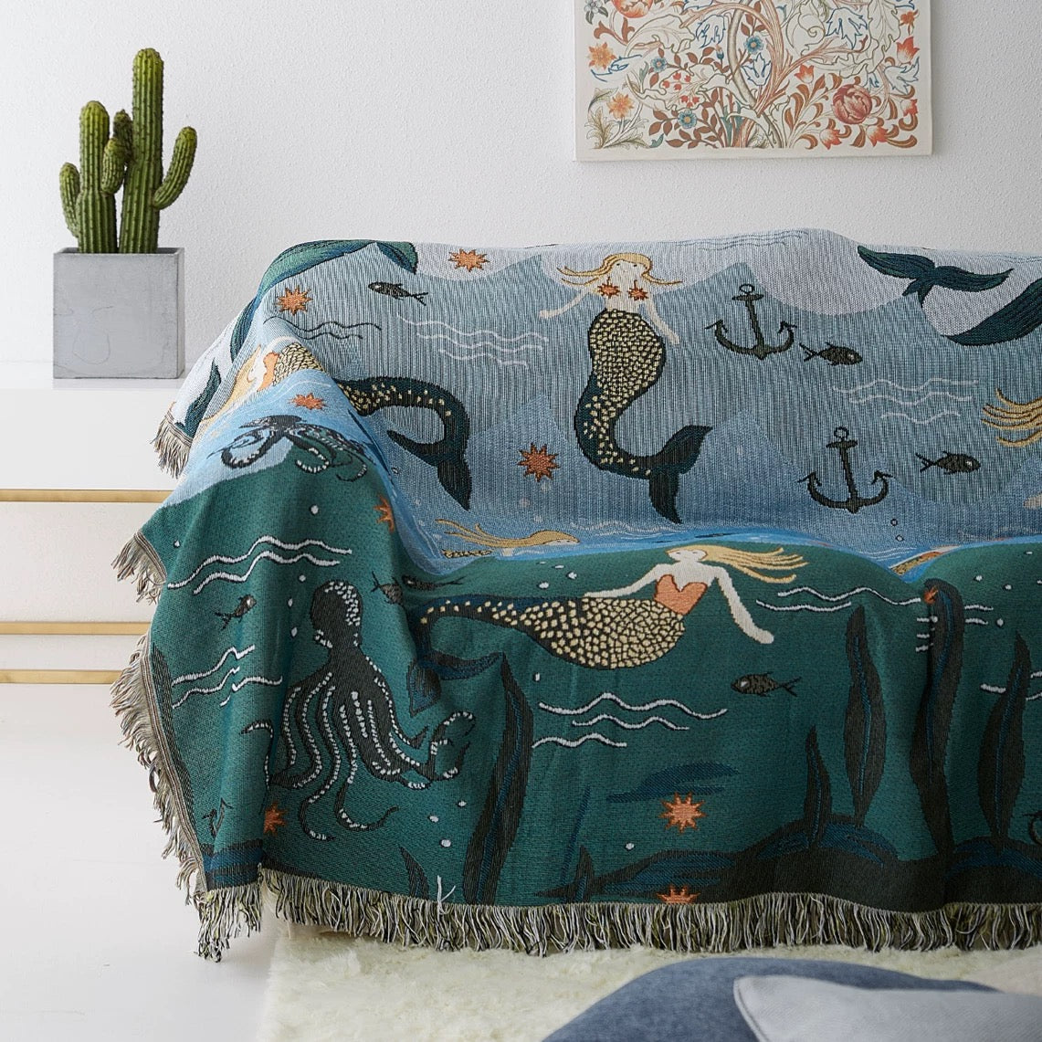 Mermaid-themed blanket draped over a chair with a cactus plant and framed artwork in the background.